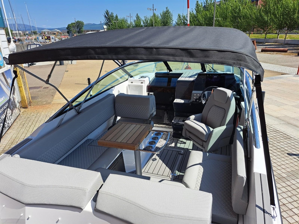 Interior of a sleek boat featuring gray cushioned seating, a wooden table, and modern control panel under a black canopy.